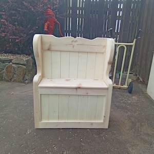 May include: A wooden storage bench with a high back and armrests. The bench is made from light-coloured wood and has a hinged seat for storage. A heart-shaped cutout decorates the backrest. The bench is outdoors.
