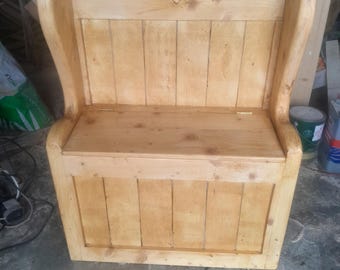 May include: A wooden storage bench with a high back and armrests. The bench is made of light-colored wood and features a carved heart detail on the backrest. The seat lifts to reveal storage space. The bench is designed for indoor use.