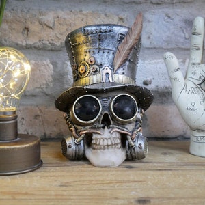 May include: A steampunk skull ornament with goggles, a top hat, and a feather. The skull has a mustache and respirator. A decorative light bulb lamp and a palmistry hand statue are also visible.