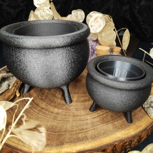 Könnte beinhalten: Zwei schwarze Kessel aus Gusseisen mit jeweils drei Beinen. Der größere Kessel ist höher und breiter als der kleinere. Beide Kessel stehen auf einer Holzoberfläche.