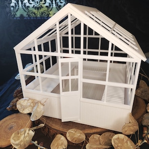 Op de afbeelding: Een witte miniatuurkas met een zadeldak en glazen wanden en deur. De binnenkant heeft een lattenbank en vloer. De structuur staat op een houten basis, omringd door gedroogde zaaddozen en kleine stenen.