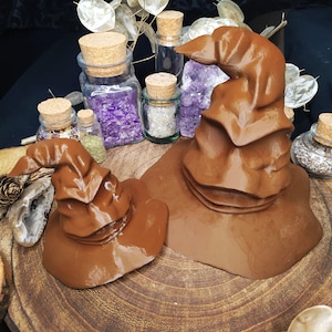 Könnte beinhalten: Zwei braune Filzhüte, einer mit einer spitzen Spitze und der andere mit einem breiten Rand, werden auf einer Holzoberfläche präsentiert. Die Hüte sind von kleinen Glasflaschen umgeben, die mit bunten Kristallen und getrockneten Blumen gefüllt sind.