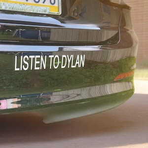May include: A black car bumper with the white text "LISTEN TO DYLAN" in a sans-serif font. The bumper has a license plate at the top and a small orange reflector on the right side.