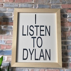 Può includere: Un cartello incorniciato con la scritta "I LISTEN TO DYLAN" in lettere nere su sfondo bianco. La cornice è di colore legno chiaro e il cartello è esposto su un muro di mattoni.