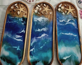 May include: Three elongated oval wooden trays featuring a resin ocean design. Each tray displays a sandy beach section with small seashells and a starfish, transitioning into deep blue and turquoise water with white foam waves.