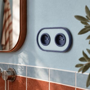 Op de afbeelding: Een dubbel elektrisch stopcontact met een donkerblauw ovaal frame en een lichtblauwe rechthoekige afdekplaat. Het stopcontact heeft twee ronde openingen met randaarde en is gemonteerd op een lichtblauwe muur boven een betegelde achterwand.
