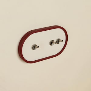 Peut inclure: Une plaque d'interrupteur murale de forme ovale avec un panneau central blanc encadré par une bordure rouge foncé. La plaque comprend trois interrupteurs à bascule métalliques, disposés avec un à gauche et deux rapprochés à droite.