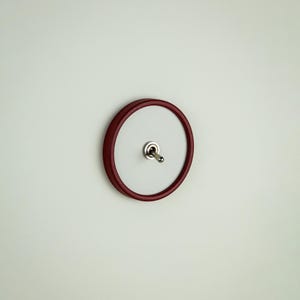 Puede incluir: Un interruptor basculante redondo rojo con una palanca plateada. El interruptor está montado en una placa circular blanca, sobre una pared blanca lisa. El diseño es clásico y minimalista.