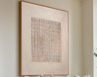 May include: A large square framed wall art piece featuring a textured beige grid pattern on a light cream background. The artwork is displayed on a plain wall above a white sofa with beige throw pillows and a wooden coffee table in a bright room.