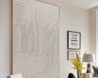 May include: Large abstract textured wall art in neutral tones. The artwork features a series of curved and linear patterns in a cream color, set within a gold frame. The piece is displayed in a modern living room setting.