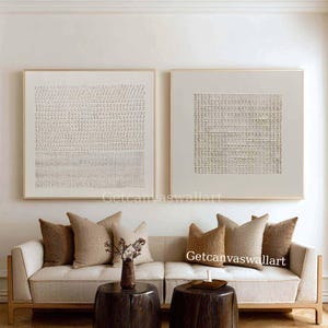 Puede incluir: Dos cuadros cuadrados enmarcados con patrones abstractos texturizados en beige con motivos de rejilla y puntos. Las obras cuelgan sobre un sofá de color claro con cojines marrones y dos mesas auxiliares cilíndricas de madera oscura.