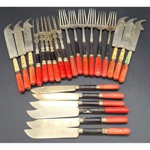 May include: A collection of antique silverware, including knives, forks, and cheese knives. The handles are a mix of red, black, and cream colors. The blades and tines are silver-toned, and the set is arranged on a dark surface.