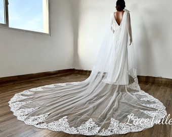 May include: A white wedding dress with a long train and a sheer cape. The cape is attached at the shoulders and drapes down the back. The train and cape feature intricate white lace detailing. The image is set in a room with a window.