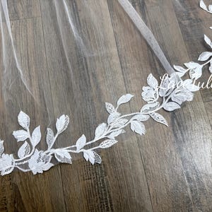 Peut inclure: Un voile de mariée blanc transparent avec une délicate bordure en dentelle florale. La dentelle présente des feuilles et des fleurs blanches détaillées, créant un design élégant et romantique. Le voile est présenté sur une surface en bois.