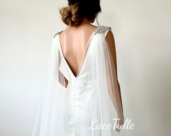 May include: A white bridal gown with a low back and a sheer tulle cape. The shoulders of the gown are embellished with sparkling crystals. The cape flows down the back, creating an elegant and ethereal look. The text "Lace Tulle" is visible at the bottom.