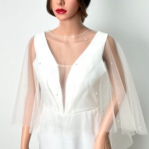 May include: White bridal cape with a sheer tulle overlay and pearl embellishments. The cape has a V-neckline and is sleeveless.