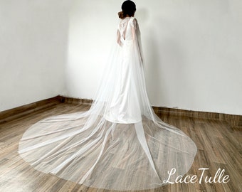 May include: A white tulle wedding veil with a long train. The veil is draped over a white dress and the model is standing on a wooden floor.