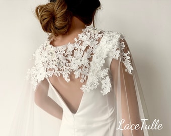 May include: White lace bridal cape with a delicate floral design and a sheer tulle overlay. The cape is worn over a white dress with a low back.
