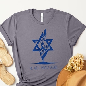May include: A gray t-shirt with a blue Star of David and a silhouette of a person dancing. The text "We will dance again" is printed below the design.