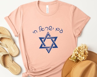 May include: A light pink t-shirt featuring a blue Star of David and Hebrew text. The shirt is styled with a pair of woven sandals and a brown hat. The shirt is made of a soft material.