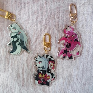 Puede incluir: Tres llaveros con personajes de dibujos animados de la serie animada Helluva Boss. El primer llavero presenta un lobo blanco con marcas negras y una corbata de lazo roja. El segundo llavero presenta un demonio de pelo rosa con una estrella negra y una corbata de lazo roja. El tercer llavero presenta un lobo blanco con marcas negras y una corbata de lazo roja, junto con un demonio rojo y negro con cuernos.