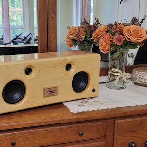Può includere: Un altoparlante rettangolare in legno con due altoparlanti neri e due fori più piccoli. L'altoparlante ha una finitura in legno chiaro e un piccolo logo sul davanti. È appoggiato su un comò di legno con fiori e candele sullo sfondo.