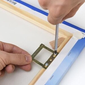 Op de afbeelding: Een persoon gebruikt een schroevendraaier om een metalen ophangsysteem aan een houten canvasframe te bevestigen. Het ophangsysteem heeft een klein, rechthoekig frame met een rasterpatroon en is ontworpen om een afbeelding of kunstwerk te houden.