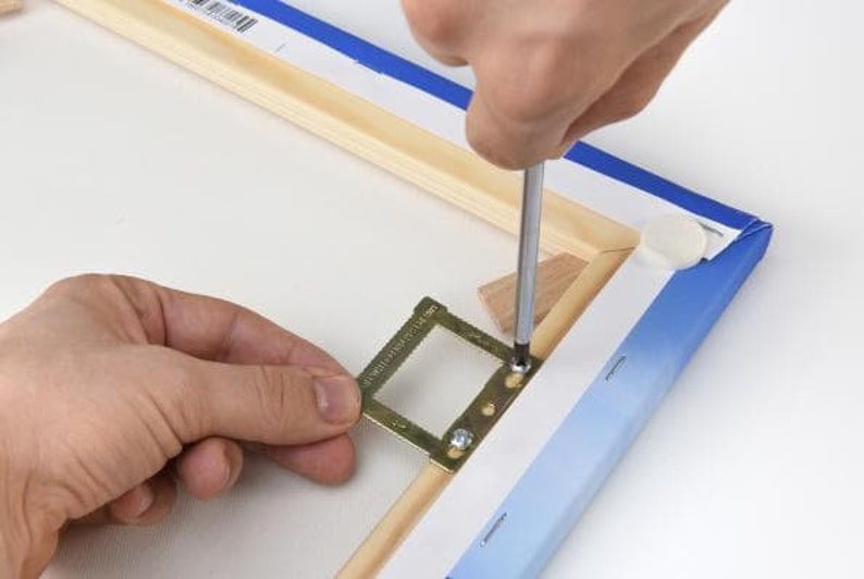 Op de afbeelding: Een persoon gebruikt een schroevendraaier om een metalen ophangsysteem aan een houten canvasframe te bevestigen. Het ophangsysteem heeft een klein, rechthoekig frame met een rasterpatroon en is ontworpen om een afbeelding of kunstwerk te houden.