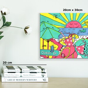 Op de afbeelding: Een kleurrijke illustratie van een strandsc&egrave;ne met een zon, een parasol, een reddingsbrigadepost, een surfplank, een drankje met een rietje en de tekst "Ocean Dr" en "Frisbee".