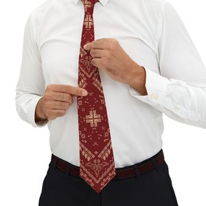 Op de afbeelding: Een bordeauxrode stropdas met een herhalend beige kruis- en geometrisch patroon. De stropdas wordt aangepast door een persoon die een wit overhemd en een zwarte broek draagt. Een klassiek accessoire.