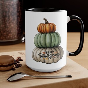 Könnte beinhalten: Weiße Keramiktasse mit schwarzem Rand und Henkel. Die Tasse zeigt einen Stapel von drei Kürbissen mit dem Text "Happy Thanksgiving" in einer dekorativen Schrift.