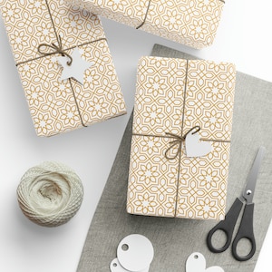 Puede incluir: Tres cajas de regalo blancas envueltas en papel con estampado geométrico dorado con cuerda marrón y etiquetas de papel blancas. Las cajas están sobre una superficie de tela gris con círculos de papel blanco y un par de tijeras negras.