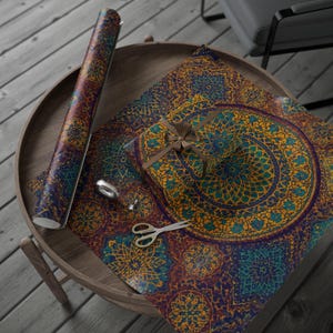 May include: A roll of patterned wrapping paper and a wrapped gift with a brown ribbon sit on a wooden table. The paper features a repeating mandala design in shades of blue, gold, and purple. A pair of scissors and a silver ring are also on the table.