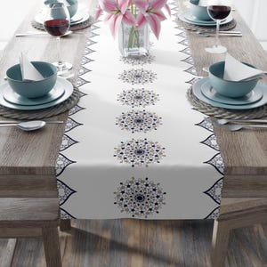 May include: White table runner with a geometric pattern in black and gold. The pattern features repeating circular designs with intricate details. The runner is laid on a wooden table with place settings.