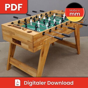 May include: A wooden foosball table with a green playing surface and black and white players. The table has six rods with black handles. The table is on a light brown wooden base with four legs. The table is labeled "Maße in mm" in German.
