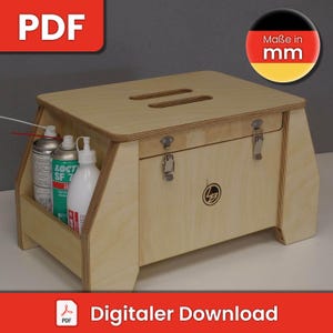 Puede incluir: Una caja de almacenamiento de madera con una tapa con bisagras y un pequeño estante. La caja está hecha de madera contrachapada y tiene un acabado de madera clara. La tapa tiene dos recortes para transportar. La caja está sentada sobre una superficie gris. El texto "Maße in mm" es visible en una pegatina roja y amarilla.