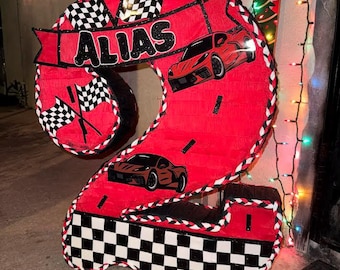 May include: A red number two shaped piñata featuring a black and white checkered flag pattern at the base and top. The piñata displays the name ALIAS in black letters on a red banner, accompanied by two illustrations of red sports cars.