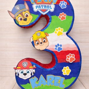 Puede incluir: Una piñata número tres colorida con personajes de Paw Patrol. La piñata está decorada con rayas azules, rojas y verdes, huellas de patas y el nombre "KAREL". El logo de Paw Patrol está en la parte superior.