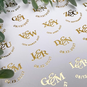Peut inclure: Autocollants de mariage dorés avec initiales et dates, comme "G&M 08.12.2027" et "J&R 08.12.2021". Les autocollants ronds sont sur une surface blanche, avec des feuilles d'eucalyptus dans le coin.