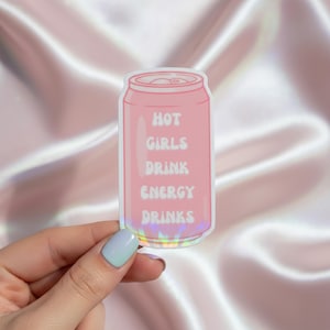 Könnte beinhalten: Ein rosa Aufkleber in Form einer Getränkedose mit den Worten "HOT GIRLS DRINK ENERGY DRINKS" in Weiß. Der Aufkleber hat einen holografischen, irisierenden Rand und wird von einer Hand mit einem hellblau lackierten Fingernagel gehalten.