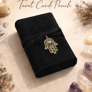 May include: Black tarot card pouch with a wrap-around tie closure and a gold-colored Hamsa hand charm. The pouch has the words "Tarot Cards Pouch" printed on the top. The pouch is made of a soft, suede-like material.