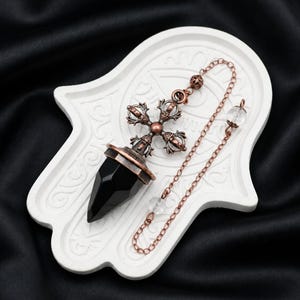 Könnte beinhalten: Ein schwarzer Obsidian-Kristall-Pendel mit einem kupferfarbenen Metallaufsatz in Kreuzform. Das Pendel ist an einer kupferfarbenen Kette mit kleinen klaren Perlen befestigt und liegt auf einer weißen, handförmigen Ablage mit geprägten Mustern.