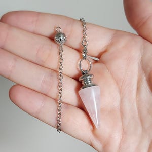 May include: A light pink, cone-shaped rose quartz pendulum attached to a silver-toned metal chain. The chain features a small, round, filigree-style metal bead at the end. The pendulum has a smooth, polished finish and a silver-toned metal cap.