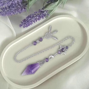 Pode incluir: Um pêndulo prateado com um cristal de ametista roxa facetado e uma corrente com contas roxas e um pingente de estrela. O pêndulo é exibido em uma bandeja oval branca com raminhos de lavanda ao fundo.