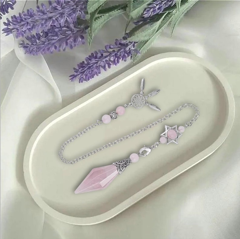 K&ouml;nnte beinhalten: Ein Rosenquarz-Pendel mit einer silbernen Kette und Akzenten, das auf einem cremefarbenen Tablett liegt. Das Pendel hat einen spitzen Kristall, einen sternf&ouml;rmigen Anh&auml;nger und rosa Perlen. Lavendelzweige sind im Hintergrund.
