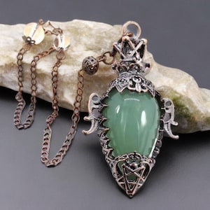 Könnte beinhalten: Eine Anhängerkette mit einem glatten, tropfenförmigen hellgrünen Stein, eingefasst in einen verzierten, dunkelmetallischen Rahmen mit filigranen Details und einem Pentagramm-Symbol an der Basis, befestigt an einer dünnen kupferfarbenen Kette mit klaren Perlen.