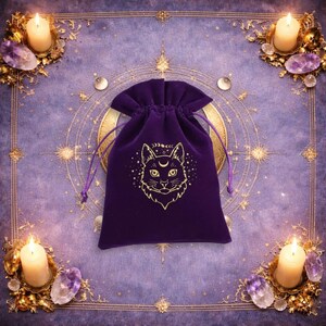 May include: A deep purple velvet drawstring pouch featuring a gold-coloured graphic of a cat face with a crescent moon on its forehead, surrounded by small stars and moon phases, with matching purple cords for closure at the top of the bag.