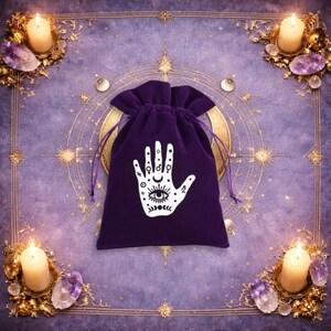 May include: A deep purple velvet drawstring pouch featuring a white graphic print of an open palm with an eye at the centre, surrounded by various celestial symbols including stars, circles, and crescent moon phases on the front surface.