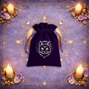May include: A deep purple velvet drawstring pouch featuring a white line art graphic of a cat face with a crescent moon on its forehead, surrounded by small stars and a lunar phase cycle above the cat's ears, with matching purple drawstrings.