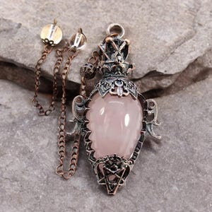 May include: A pendant necklace featuring a pink gemstone encased in an ornate, silver-toned metal frame. The pendant has a teardrop shape with decorative filigree and a star design at the bottom. A chain with two small charms and a chain is attached.
